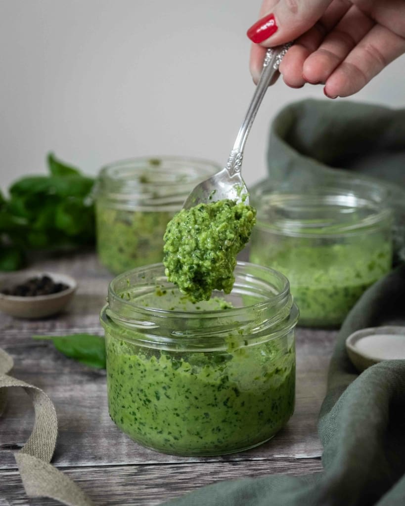 A glass bowl of vibrant green vegan pesto sat on a rustic wooden table top, with fresh basil leaves scattered around. 