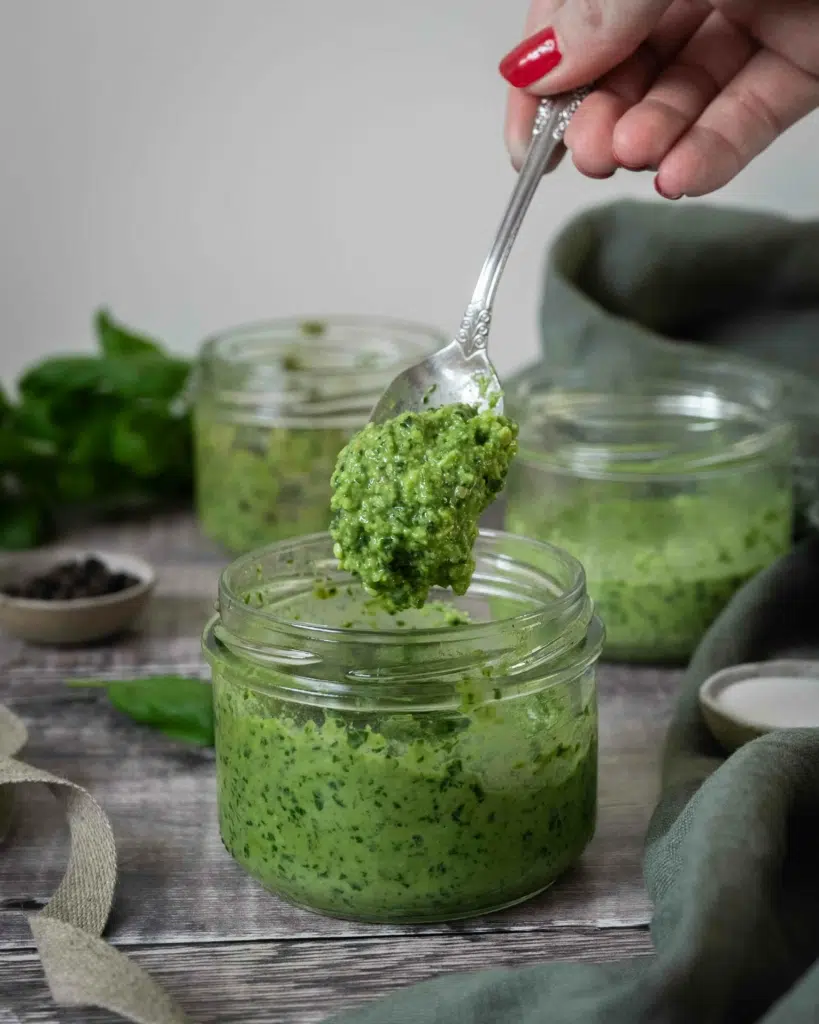 A glass bowl of vibrant green vegan pesto sat on a rustic wooden table top, with fresh basil leaves scattered around. Two bowls of pesto sit in the background - one made without pine nuts, the other is nut free.