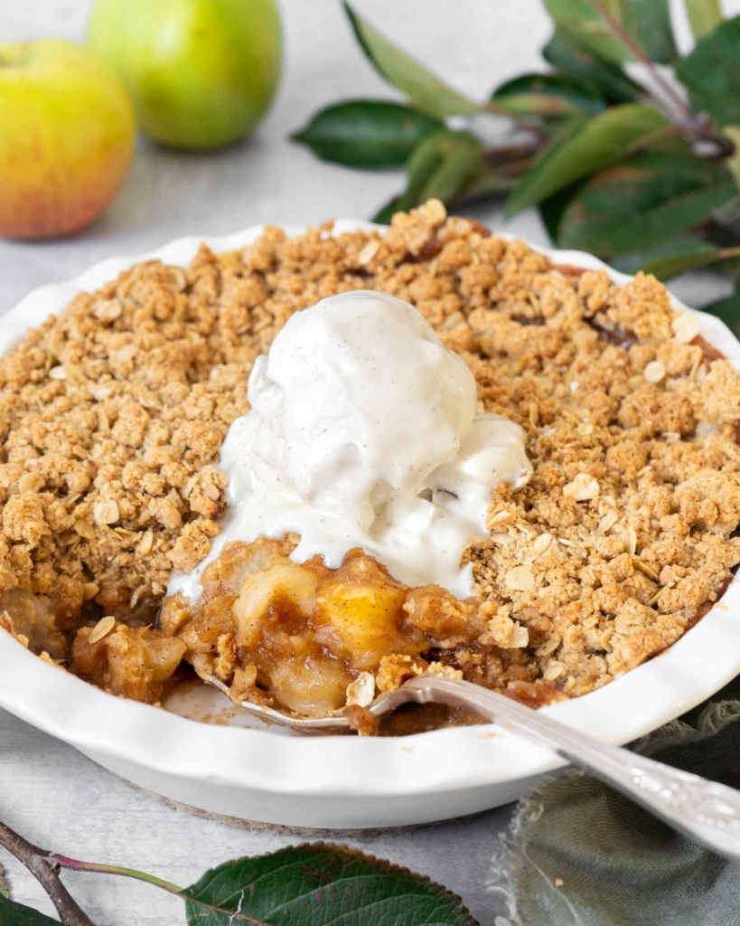 A white pie ceramic pie dish filled with vegan apple crumble no flour, with a crispy streusel topping and topped with melting vegan vanilla ice cream.