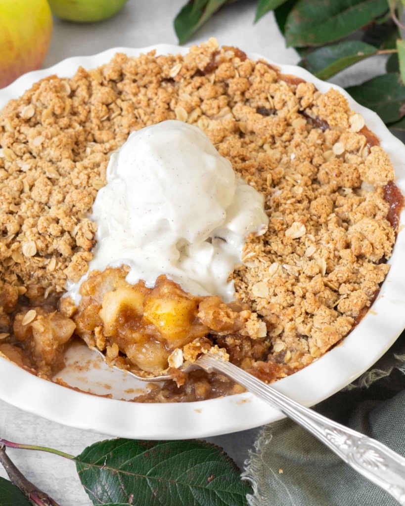 A close up photograph of an flourless apple crumble, showing the crispy streusel topping, sticky apple filling and a big scoop of vegan vanilla ice cream melting into it.