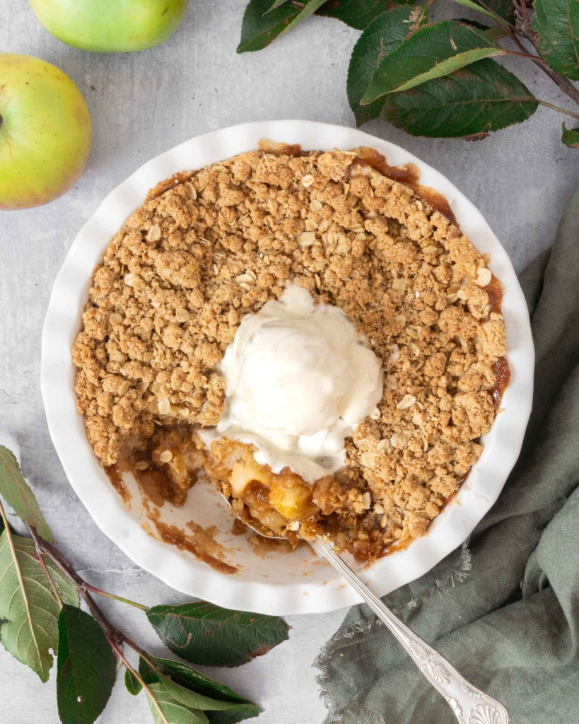 A white pie ceramic pie dish filled with vegan apple crumble, with a crispy streusel topping and topped with melting vegan vanilla ice cream.