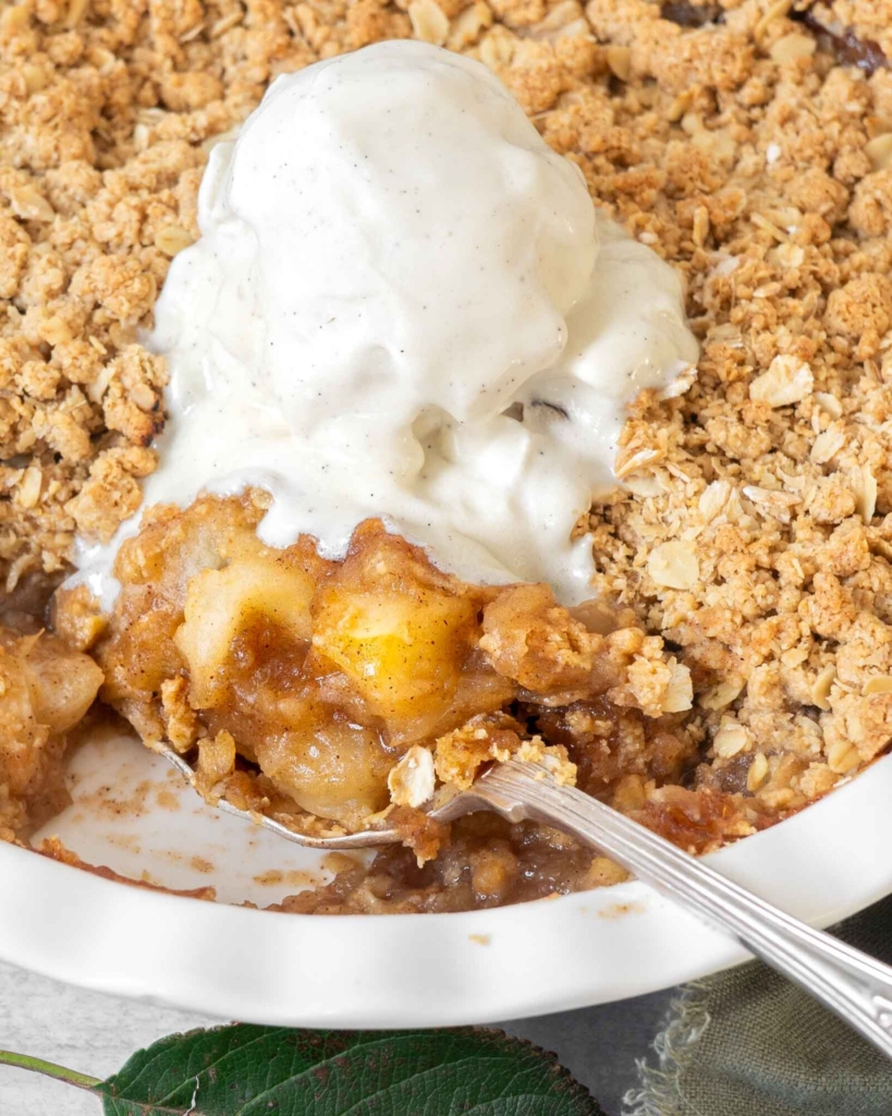 A close up photograph of an apple crumble, showing the crispy streusel topping, sticky apple filling and a big scoop of vegan vanilla ice cream melting into it.