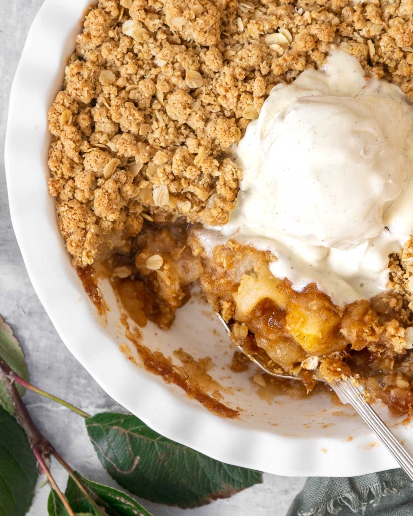 A dish of golden, crunchy apple crumble with oats and a scoop of vegan ice cream melting on top