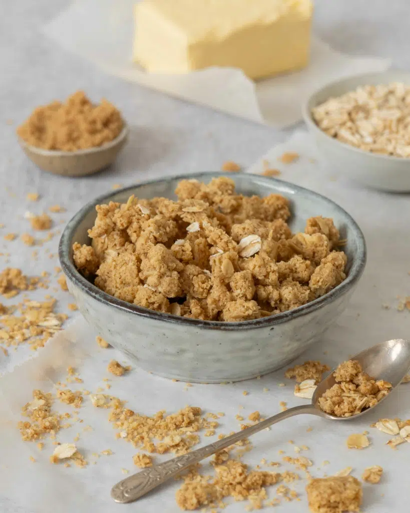 A side shot of a small bowl of crunchy streusel-style topping for crumble. In the background you can see a bowl of oats, brown sugar and a block of vegan butter.