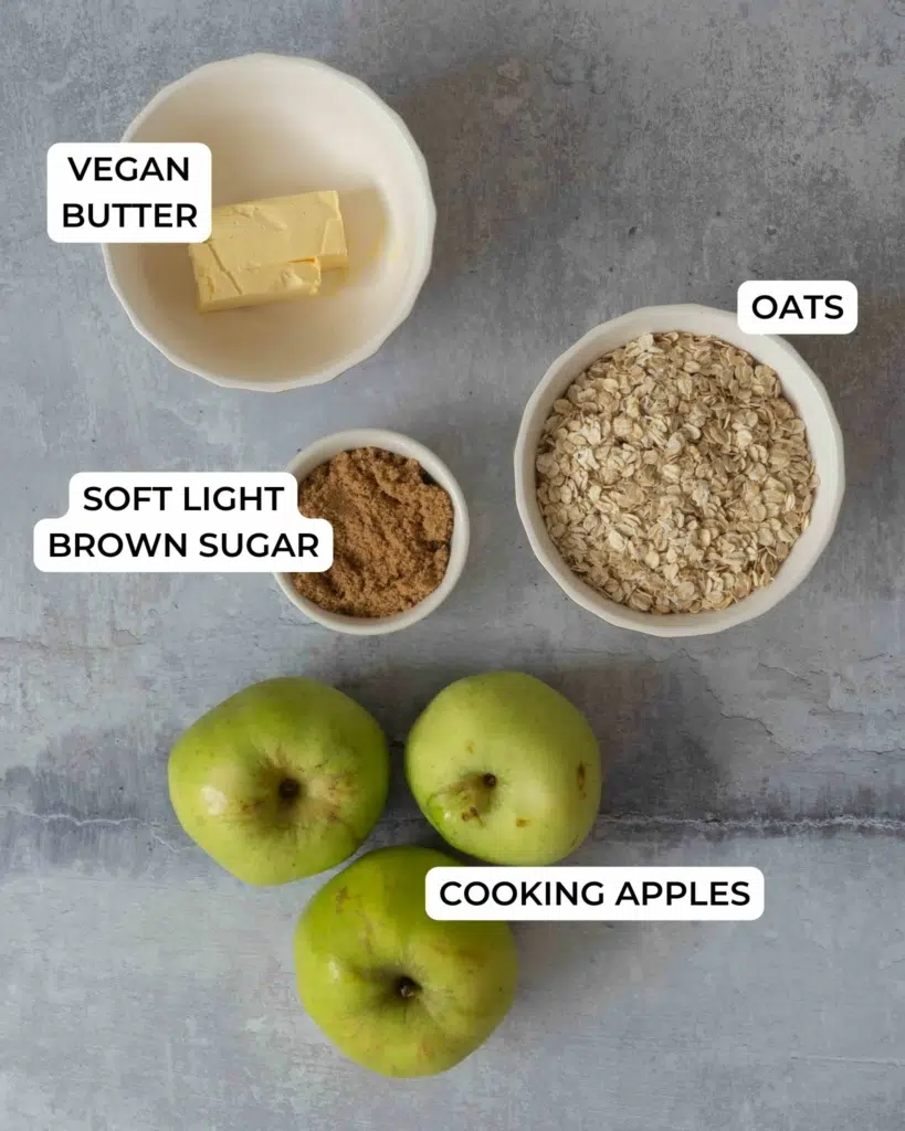 The ingredients to make a vegan apple crumble recipe laid out on a table top - apples, oats, brown sugar and vegan butter.