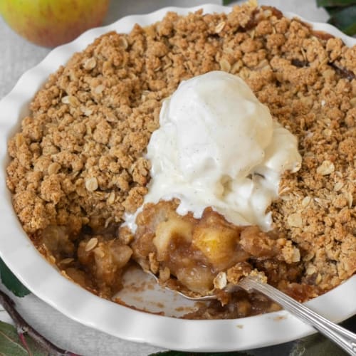 A white pie ceramic pie dish filled with vegan apple crumble with a crispy streusel topping and topped with melting vegan vanilla ice cream.