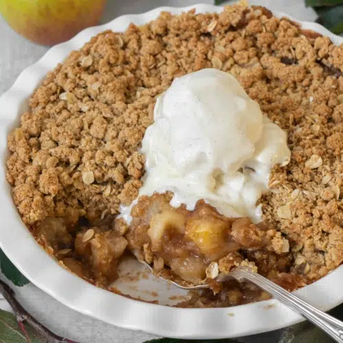 A white pie ceramic pie dish filled with vegan apple crumble with a crispy streusel topping and topped with melting vegan vanilla ice cream.