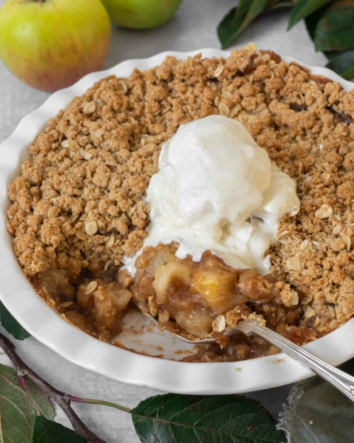 A white pie ceramic pie dish filled with vegan apple crumble with a crispy streusel topping and topped with melting vegan vanilla ice cream.