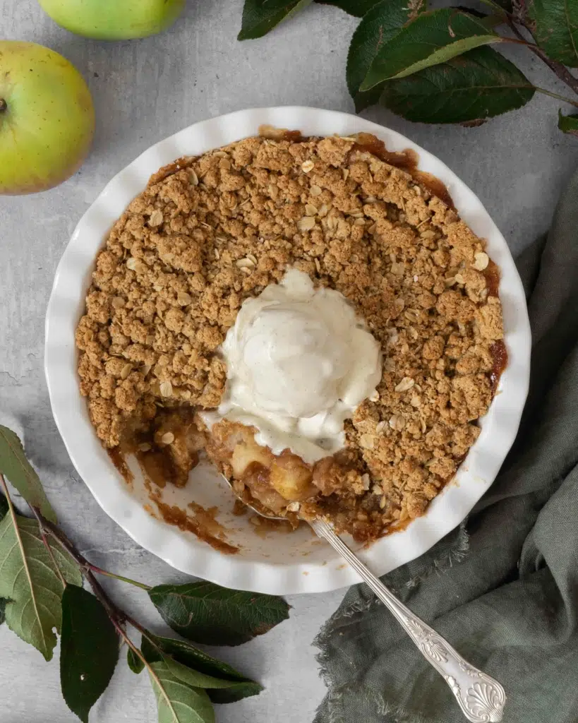 An overhead photo of an apple crumble in a round white pie dish, topped with melting vegan ice cream. Surrounded by fresh apples and branches from an apple tree.