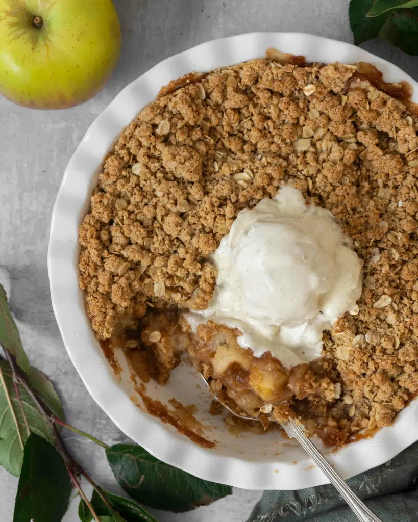 A close up overhead photo of an apple crumble in a round white pie dish, topped with melting vegan ice cream. 