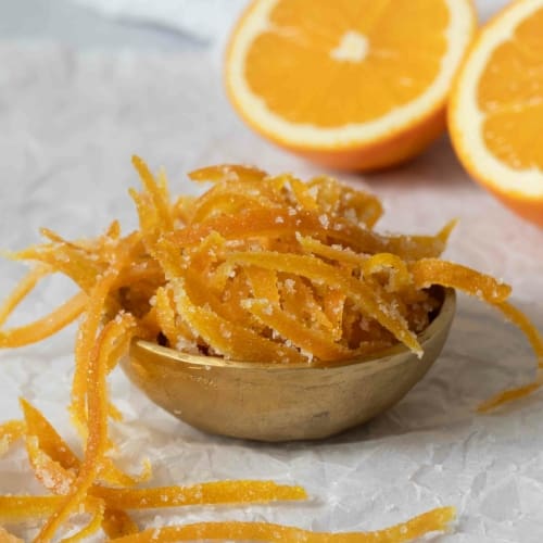 An overflowing bowl of candied orange peel, with fresh oranges in the background