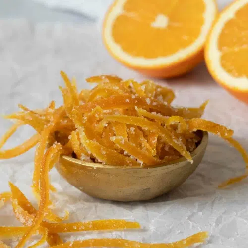 An overflowing bowl of candied orange peel, with fresh oranges in the background