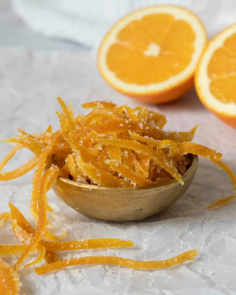 An overflowing bowl of candied orange peel, with fresh oranges in the background