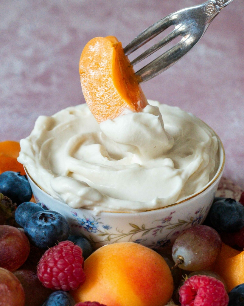 A photograph to accompany a vegan fruit dip recipe, showing a plate of colourful fruit and a vintage bowl filled with whipped vegan fruit dip. A person is dipping a piece of fresh apricot in the marshmallow fruit dip.