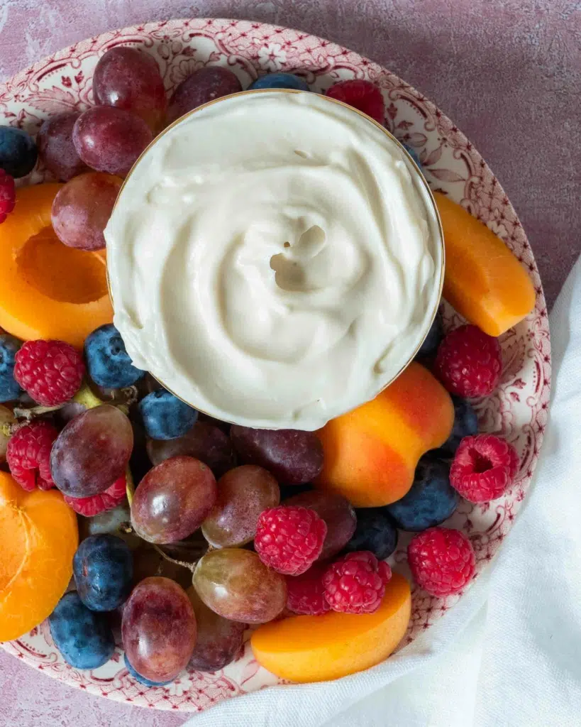 An overhead photograph of a platter of colourful fruit including grapes, raspberries, blueberries and apricots, with a vegan marshmallow fruit dip