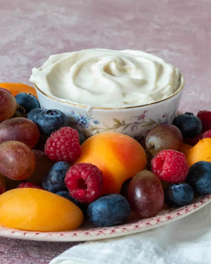 A side on photograph of a plate of colour fresh fruit and a bowl of creamy white vegan fruit dip