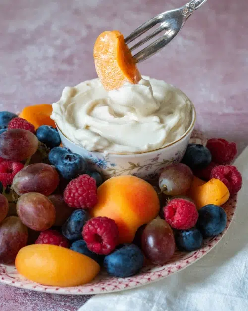 A photograph to accompany a vegan fruit dip recipe, showing a plate of colourful fruit and a vintage bowl filled with whipped vegan fruit dip. A person is dipping a piece of fresh apricot in the marshmallow fruit dip.