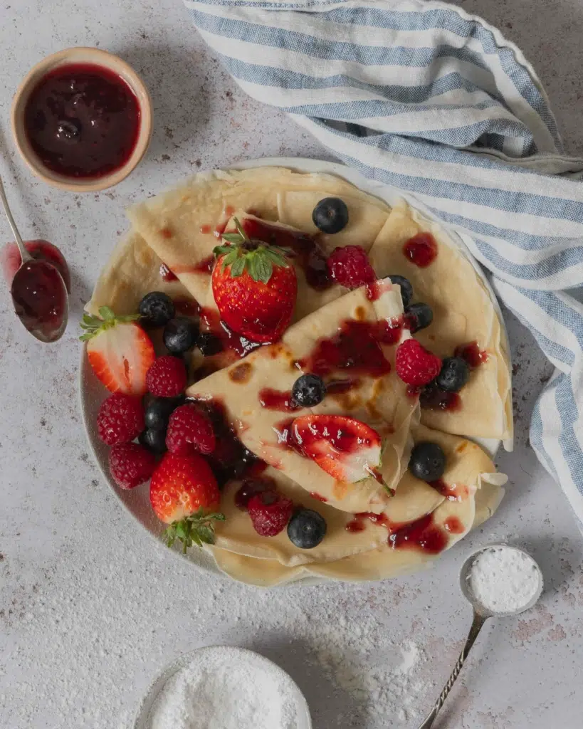An overhead photograph of vegan crepes scattered with fresh berries and drizzled with jam