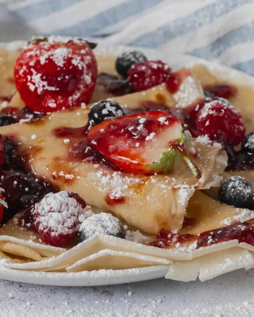 A close up photograph of a plate of vegan crepes with crisp edges and topped with fresh fruit and jam