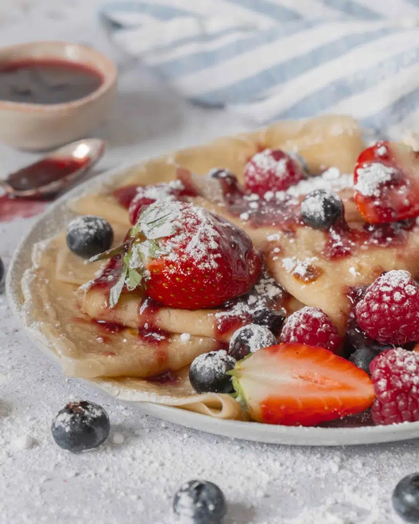 A plate of freshly cooked crepes topped with berries and icing sugar