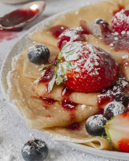 A close up photo of a plate of vegan crepes, topped with strawberries, blueberries, jam and icing sugar