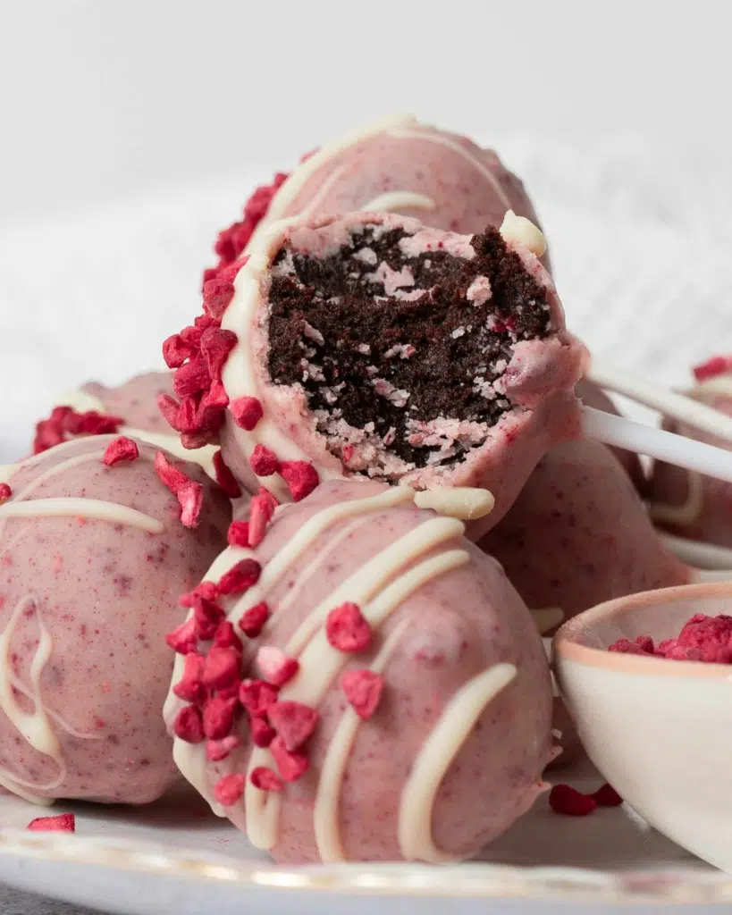 A close up of a stack of pink chocolate raspberry cake pops, one with a bite taken out revealing a dark chocolate cake interior.