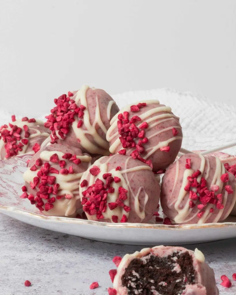 A side on photograph of a plate of speckled pink white chocolate raspberry cake pops, topped with freeze dried raspberries.