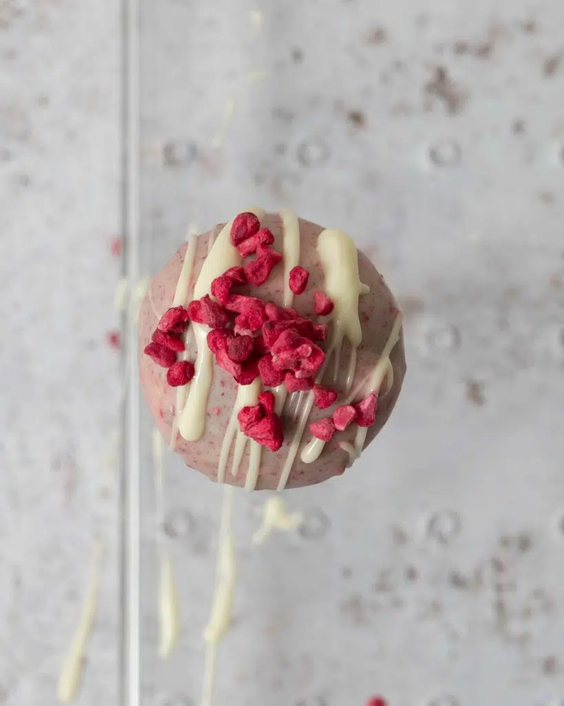 One raspberry chocolate cake pop freshly decorated with a zig zag of melted vegan white chocolate and freeze dried raspberries.