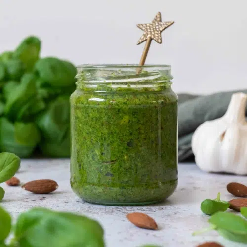 A full jar of almond pesto (vegan) surrounded by whole almonds and fresh basil leaves