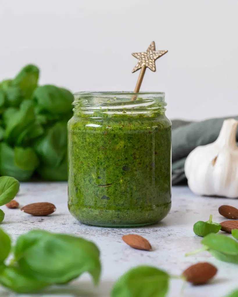 A full jar of almond pesto (vegan) surrounded by whole almonds and fresh basil leaves