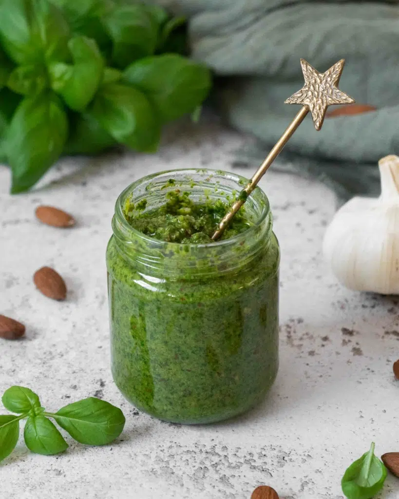 A jar of vegan almond pesto with the lid off, surrounded by fresh basil