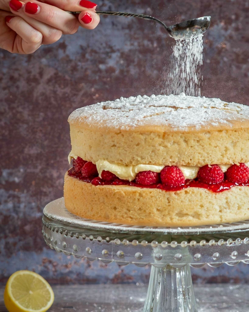A vegan lemon and raspberry cake being dusted with icing sugar