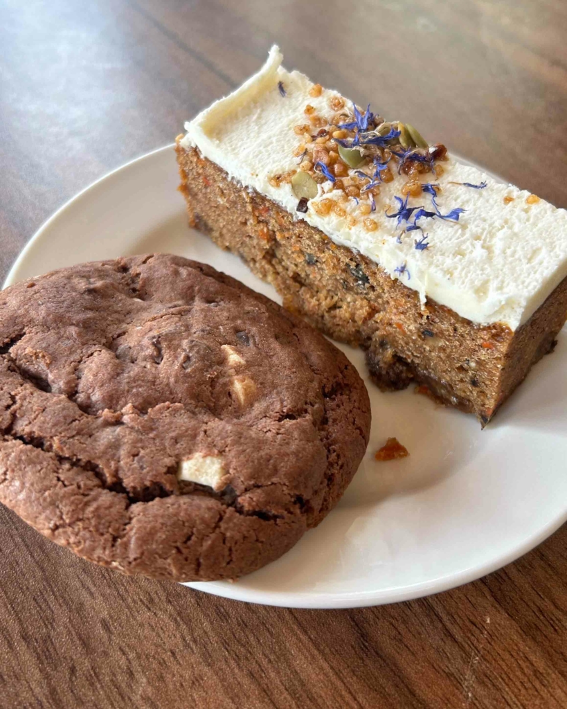 A slice of vegan carrot cake with a cream cheese frosting and a chunky chocolate chip cookie on a plate