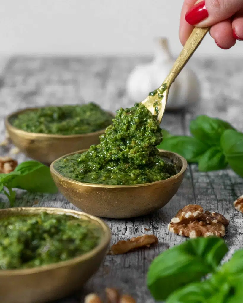 Three small golden bowls of vegan basil walnut pesto sat on a rustic table top, with a spoonful of vibrant pesto being lifted out of the middle bowl.