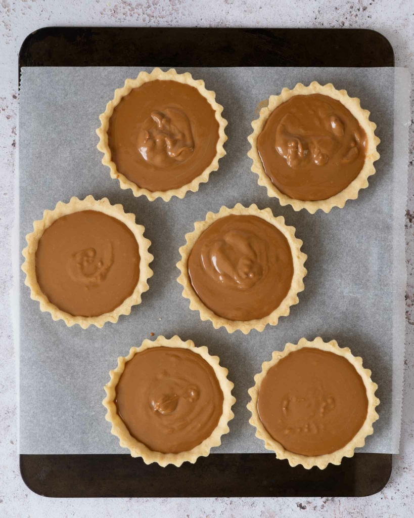 Vegan pastry tart shells on a lined tray, being filled with melted Biscoff spread