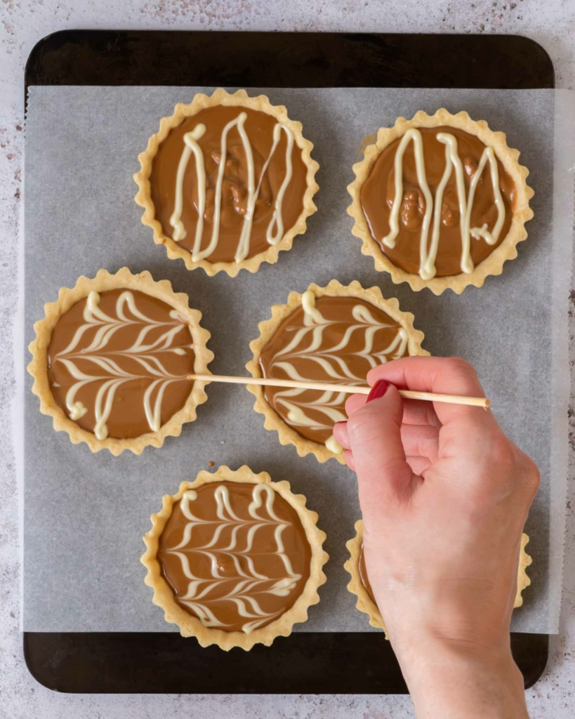 A skewer being dragged through melted white chocolate on the surface of a Biscoff tart to create a marbling effect
