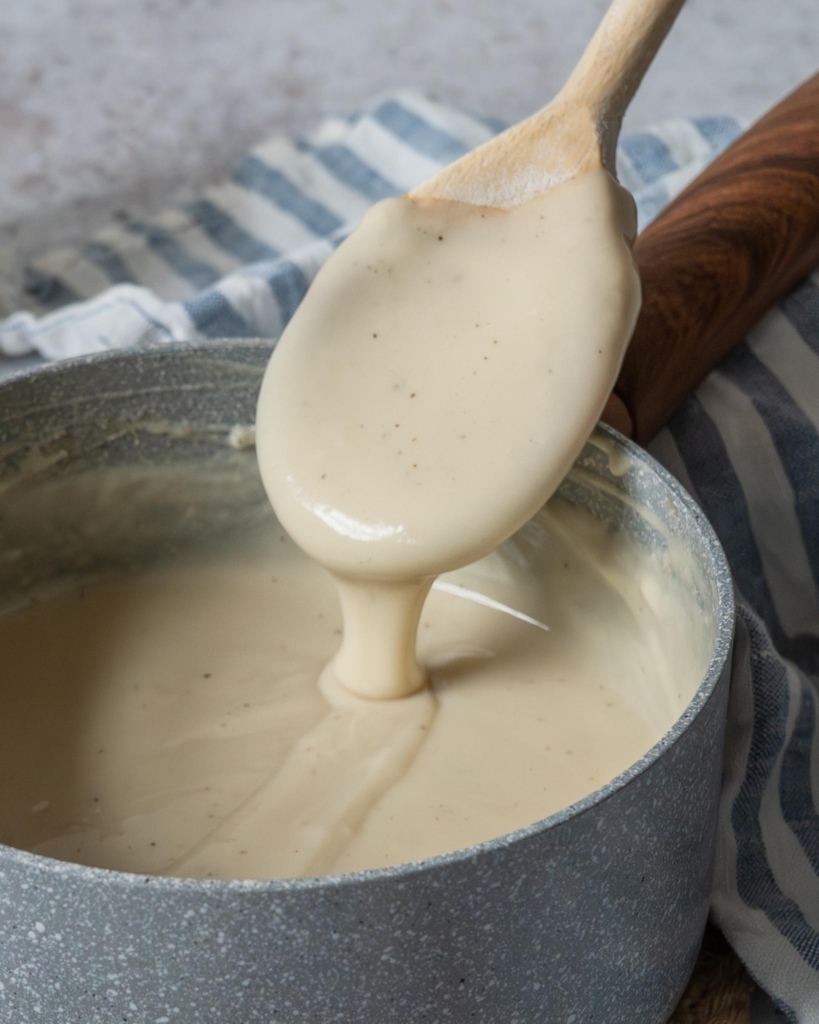 Thick and creamy vegan bechamel sauce running off of a wooden spoon into a saucepan