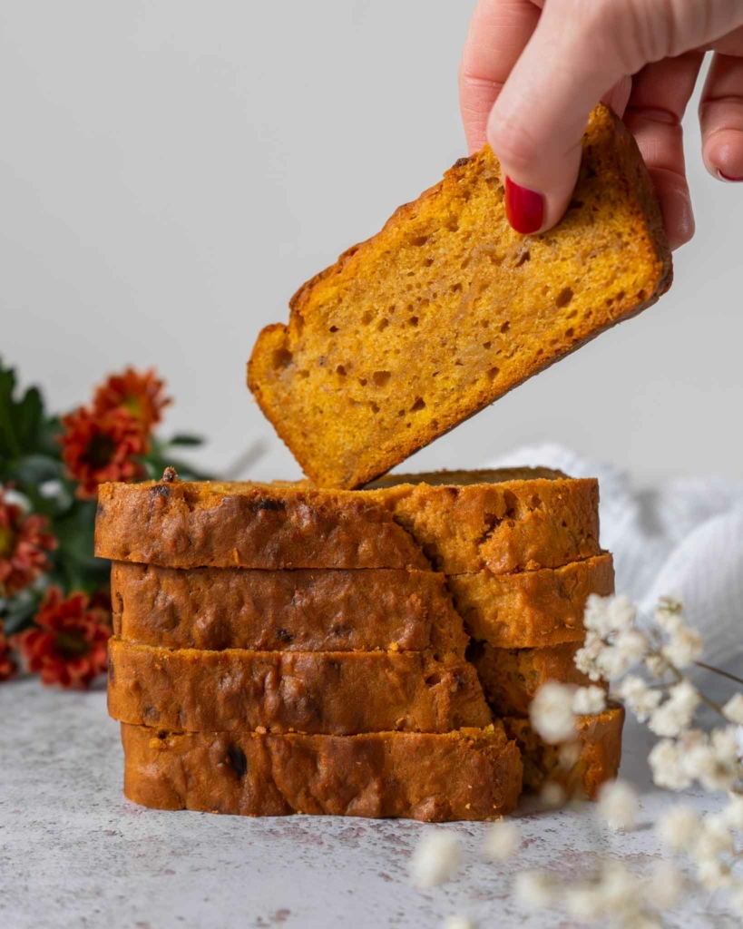 A stack of rich autumnal orange pumpkin banana bread slices on a table top, with one slice being lifted off.