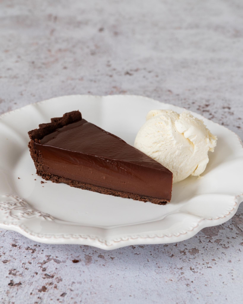 A slice of French Chocolate Tart on a white vintage plate, with a scoop of vanilla vegan ice cream beside it