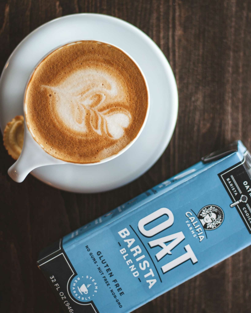 A frothy cup of coffee with a carton of Califa Farms Barista Oat Milk lying beside it