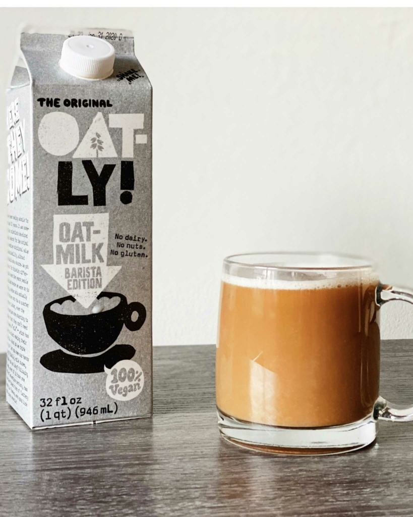 A carton of Oatly Barista oat milk and a smooth white coffee in a glass cup