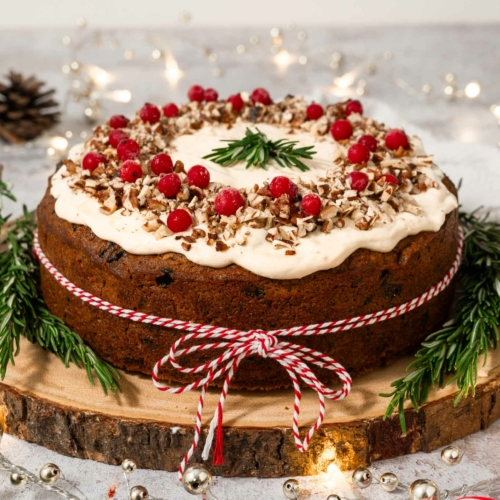 A Christmas carrot cake tied with a red and white string and decorated with fresh cranberries, rosemary sprigs and chopped nuts