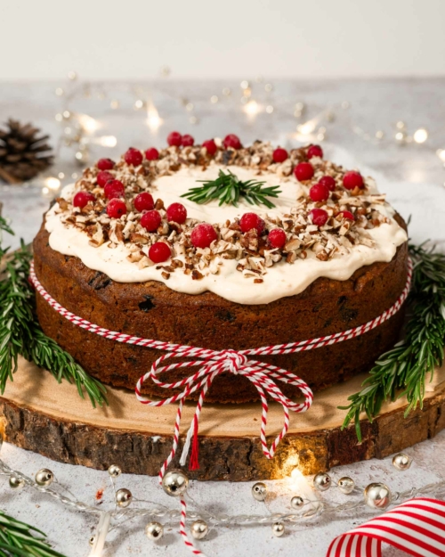 A Christmas carrot cake tied with a red and white string and decorated with fresh cranberries, rosemary sprigs and chopped nuts