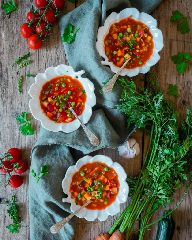 Three bowls of colourful farmhouse vegetable soup on a rustic wooden farmhouse table with fresh herbs, carrots, tomatoes and garlic scattered around