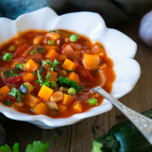A bowl of colourful farmhouse vegetable soup on a rustic wooden farmhouse table with fresh ingredients surrounding it