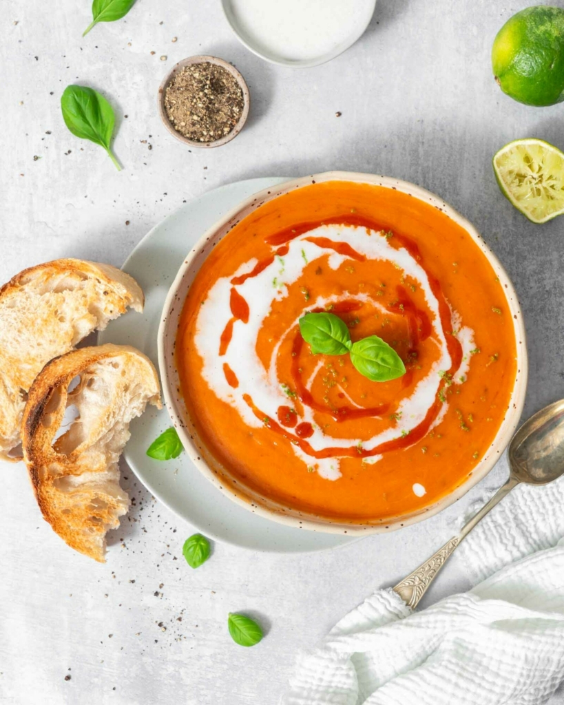 A vibrant bowl of red pepper and butternut squash soup decorated with vegan cream, fresh herbs and chilli sauce
