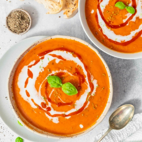 Two vibrant bowls of red pepper and butternut squash soup decorated with vegan cream, fresh herbs and chilli sauce