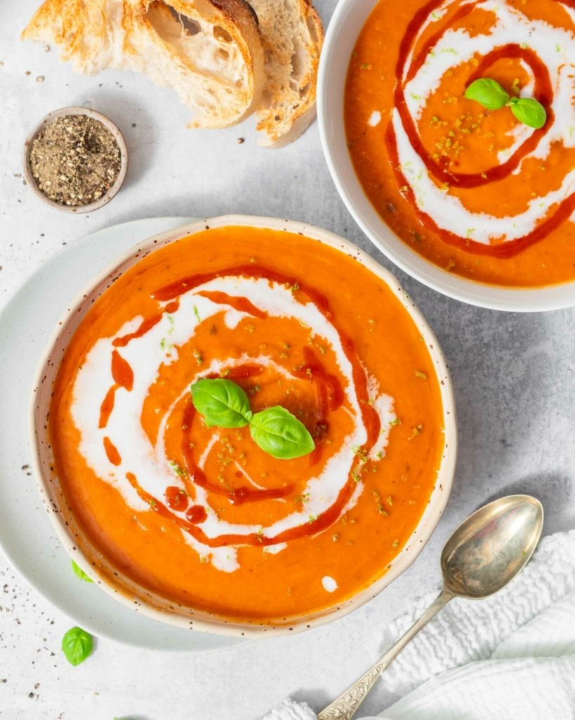 Two vibrant bowls of red pepper and butternut squash soup decorated with vegan cream, fresh herbs and chilli sauce