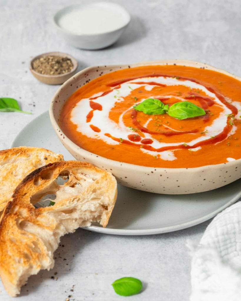 A vibrant bowl of red pepper and butternut squash soup decorated with vegan cream, fresh herbs and chilli sauce, with a side of sourdough toast