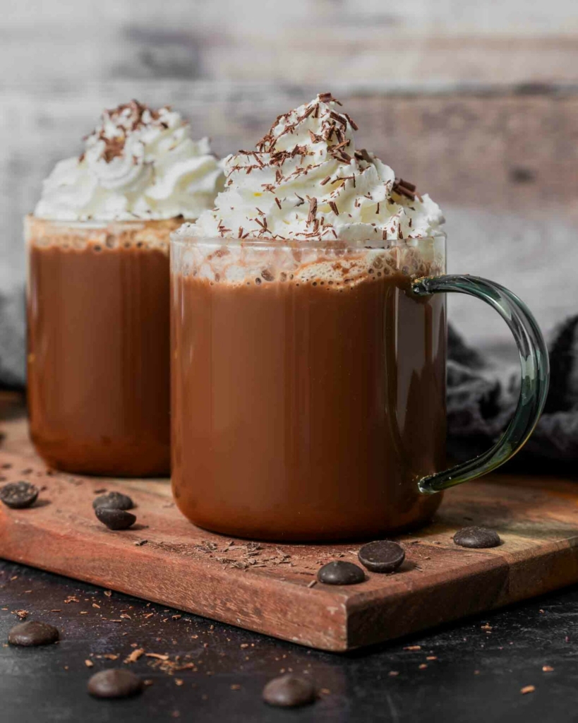 Two mugs of dairy free hot chocolate with oat milk, topped with vegan cream and grated chocolate